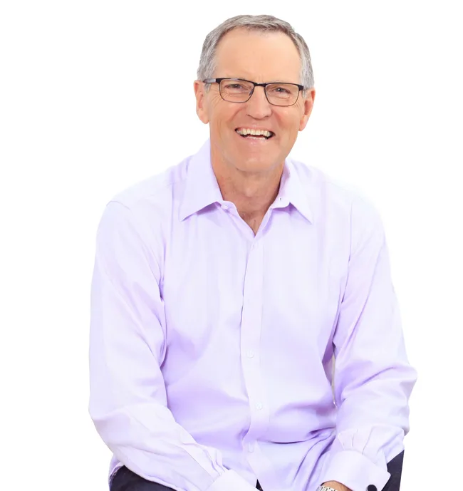 A man in glasses and a light purple shirt smiling, sitting against a white background.