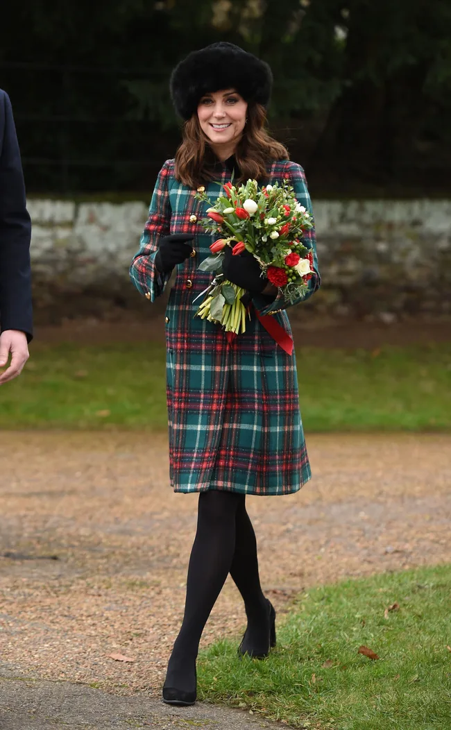 A woman in a plaid coat and black hat holds a bouquet of flowers outdoors on a grassy path.
