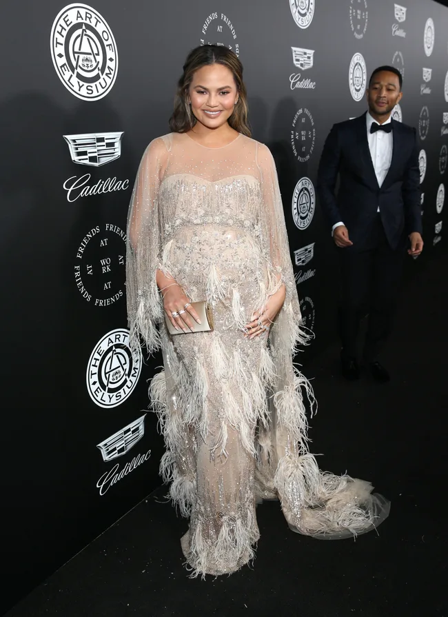 A woman in a feathered gown poses on the red carpet at The Art of Elysium event, with a man in a suit behind her.