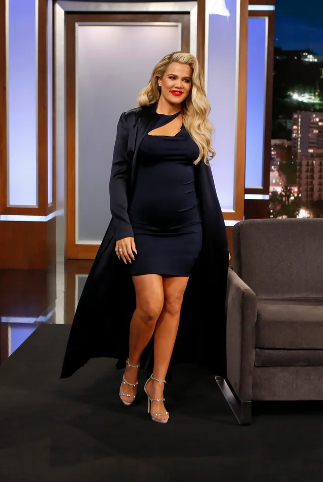 A woman in a navy dress and long coat walks on a talk show stage.