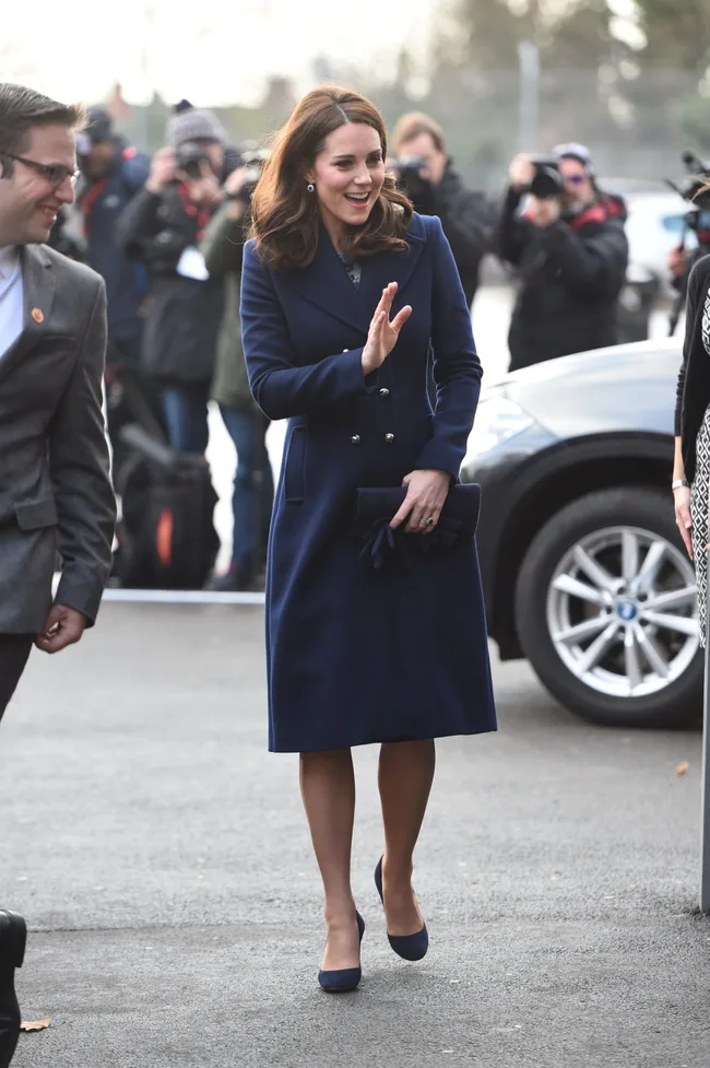 A woman in a blue coat smiles and waves while photographers capture the moment.