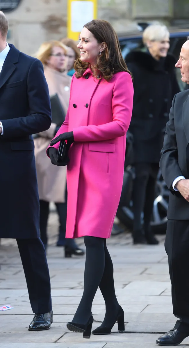 A woman in a bright pink coat smiles while standing outside with a group of people in formal attire.