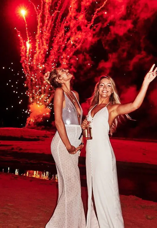 Two women in elegant dresses enjoying fireworks at night, one holding a drink and smiling, with a red-lit sky.