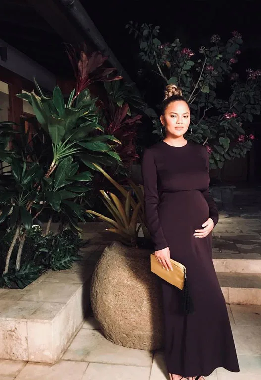 A woman in a long dark dress, cradling her belly, stands by a large potted plant at night.