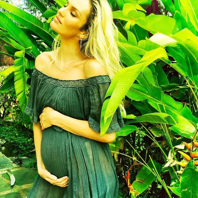 Pregnant woman in a green dress standing among lush tropical plants, gently cradling her belly.