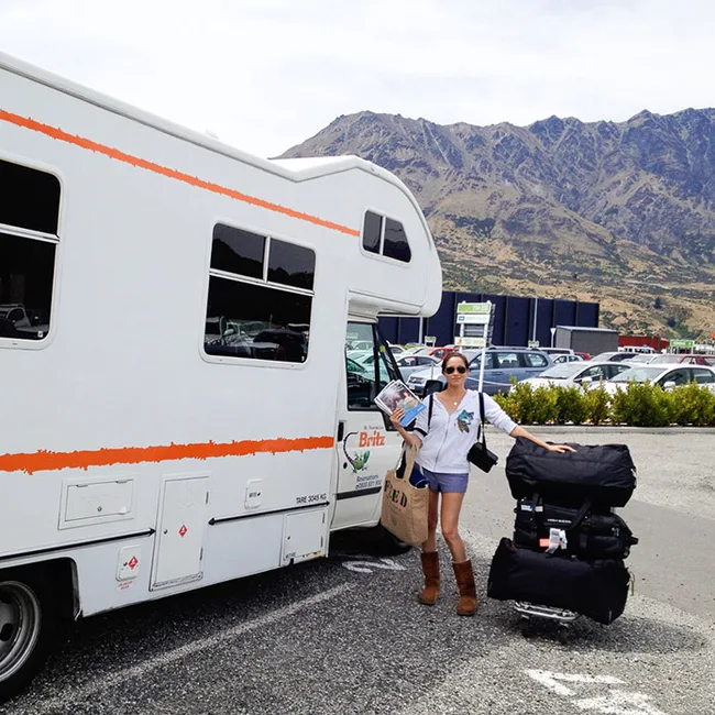 A person stands by an RV with luggage cart in a parking lot, mountainous landscape in the background.