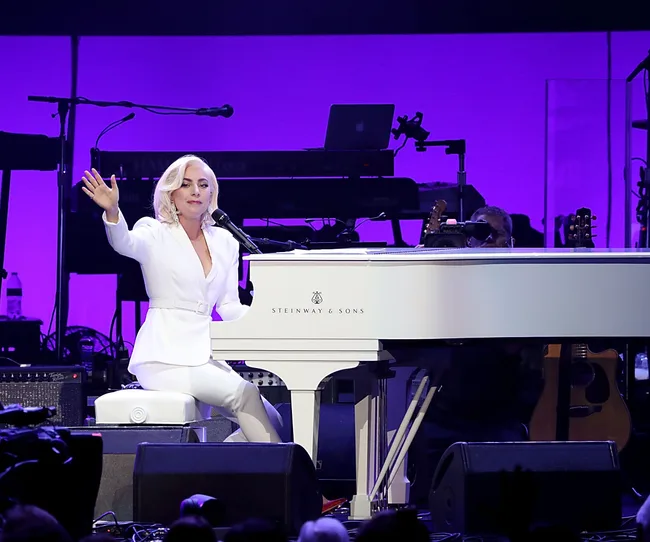 Singer in white suit performs at a white Steinway & Sons piano on stage with purple lighting.