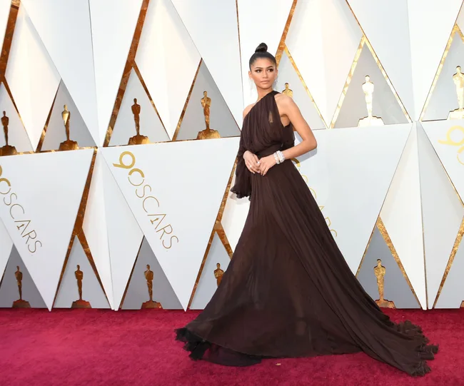 Actress in an elegant brown gown poses on the Oscars red carpet with a backdrop featuring gold statuettes.