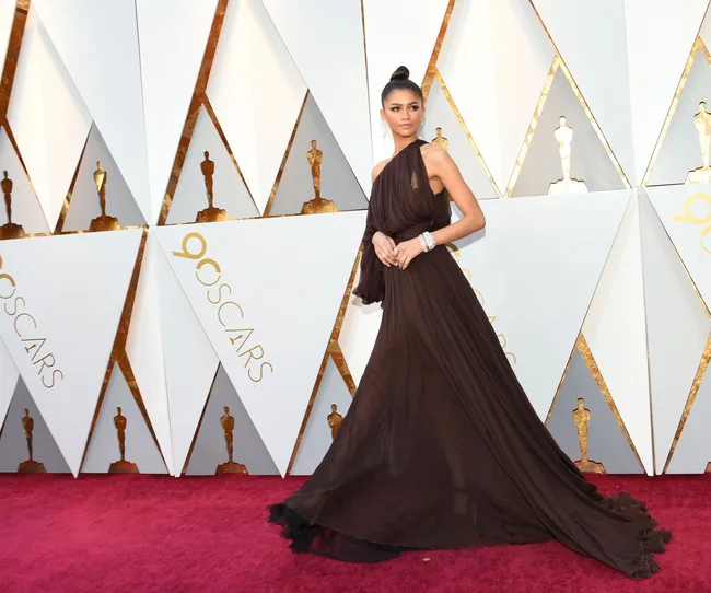 Actress in an elegant brown gown poses on the Oscars red carpet with a backdrop featuring gold statuettes.