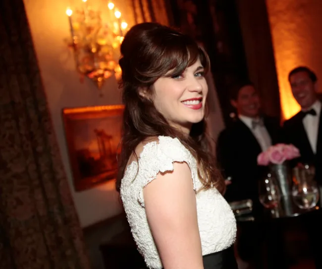 A woman in a white dress smiles at a formal event, with people and elegant decor in the background.