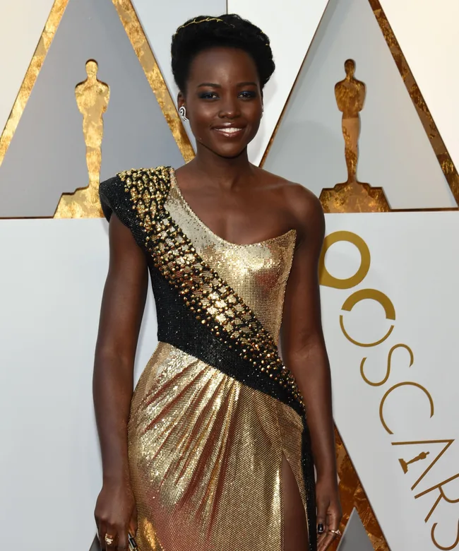 Person in a gold and black dress at an awards event with Oscar statues in the background.