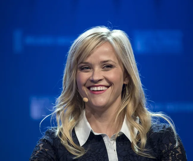Woman smiling at event with microphone, wearing black lace dress with white collar against blue backdrop.