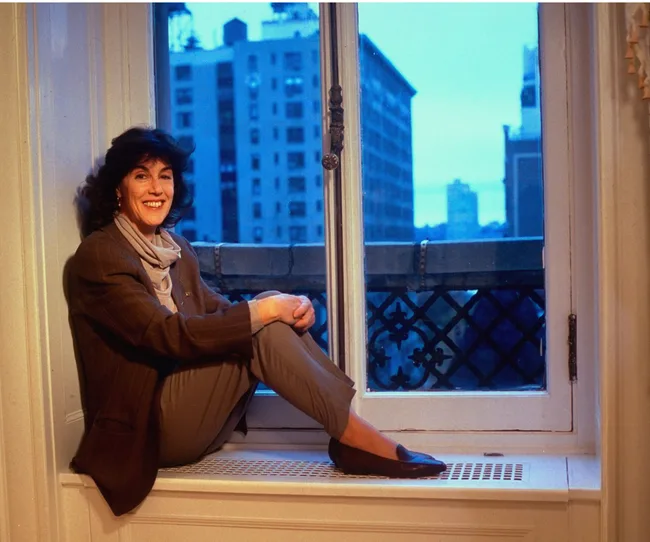 A woman smiling, sitting on a windowsill with a cityscape view at dusk.