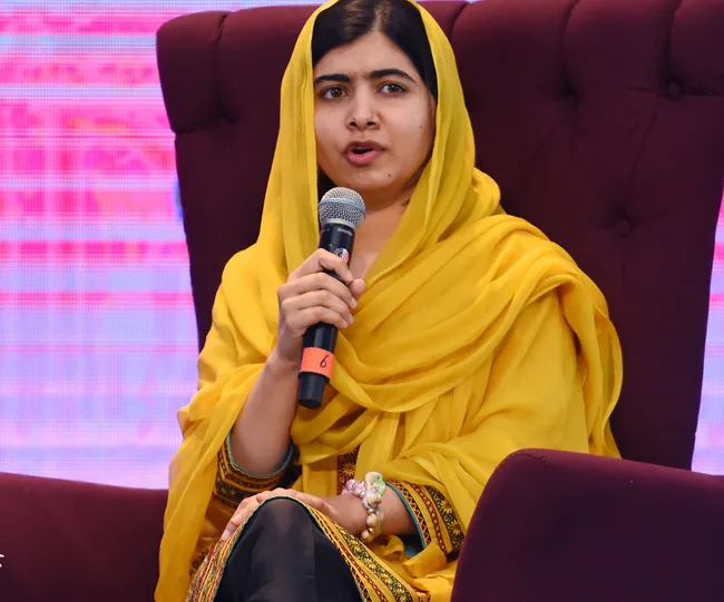 Woman in yellow headscarf and dress speaking into a microphone while seated on a stage.