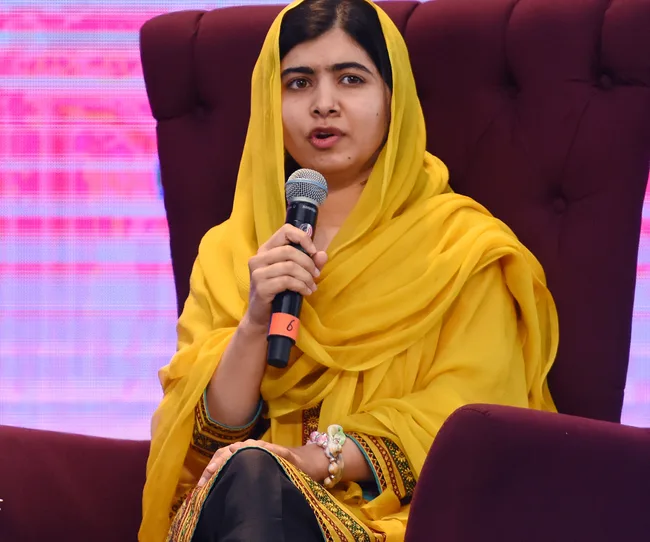 Woman in yellow headscarf and dress speaking into a microphone while seated on a stage.