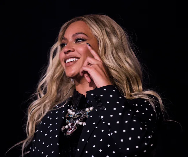 A person smiling on stage with long blonde hair, wearing a black patterned blazer and a statement necklace.