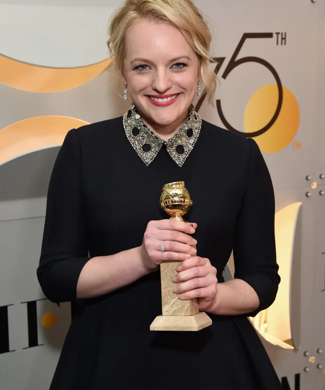 A person smiling, holding a Golden Globe award, wearing a black dress with a jeweled collar.