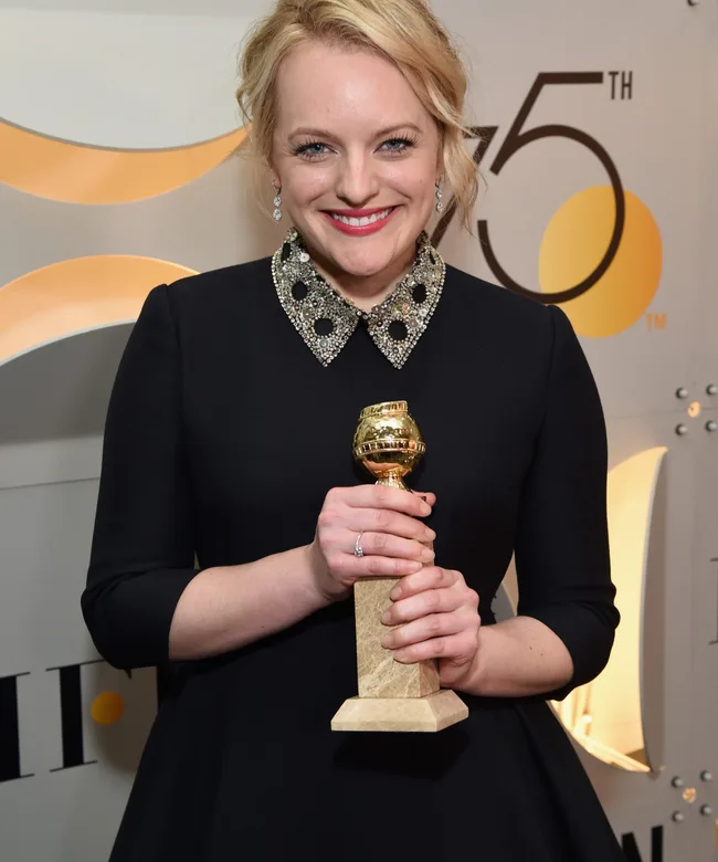 A person smiling, holding a Golden Globe award, wearing a black dress with a jeweled collar.