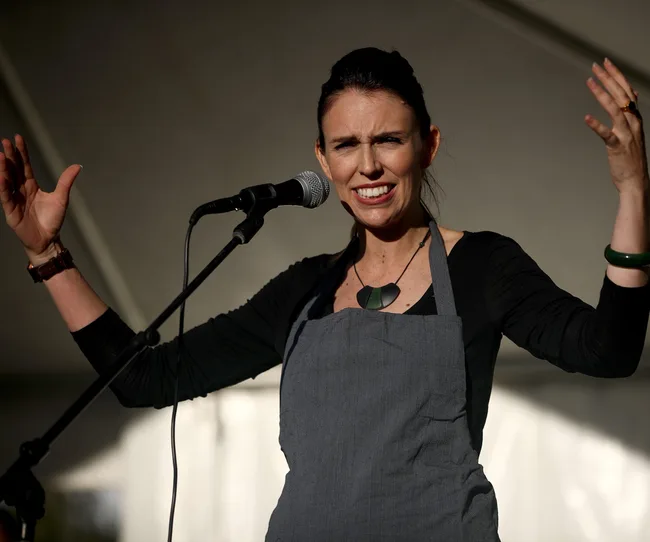 Person speaking passionately into a microphone, wearing a gray apron and dark top.