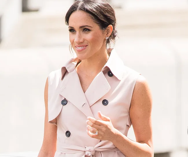 A woman wearing a sleeveless beige trench coat dress, smiling outdoors.