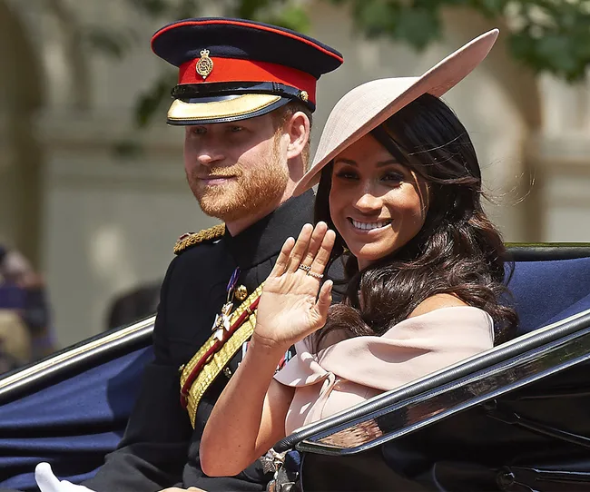 A man in military uniform and woman in a hat are riding in a carriage, with the woman smiling and waving.