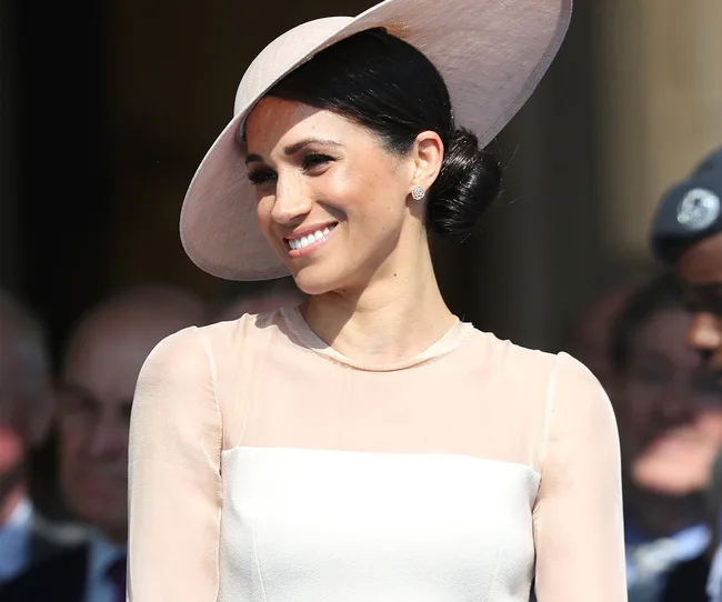 A woman in a light pink hat and dress smiling outdoors at a formal event.