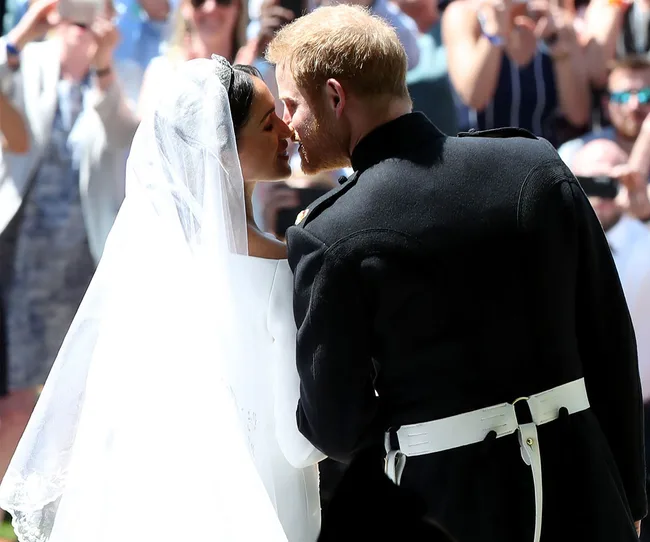 A bride and groom kiss outdoors, surrounded by onlookers, with the bride in a veil and the groom in formal attire.