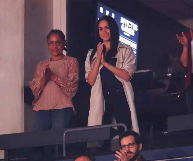 Two women standing and clapping, watching an event. Indoor setting with a screen displaying "Invictus Games" behind them.
