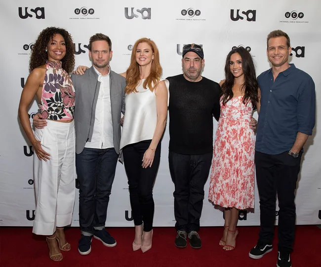 Group photo of six cast members from "Suits" on a red carpet, smiling in front of a USA Network backdrop.