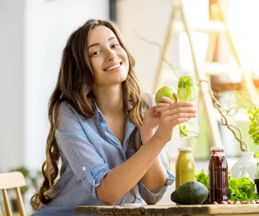 Happy woman sits at table with green vegetables and juices