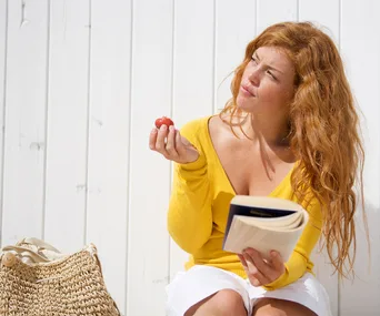 Red head young woman sitting on bench reading a book and eating a strawberry thoughful
