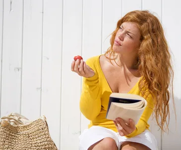 Red head young woman sitting on bench reading a book and eating a strawberry thoughful
