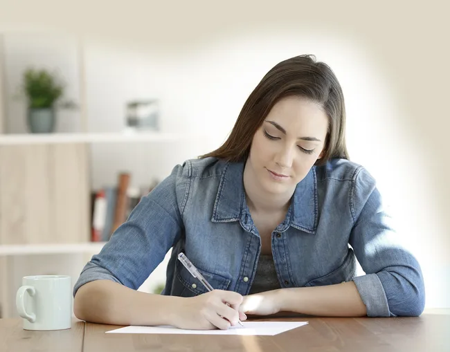 Woman in denim jacket writing at a desk with a white mug nearby in a home setting.