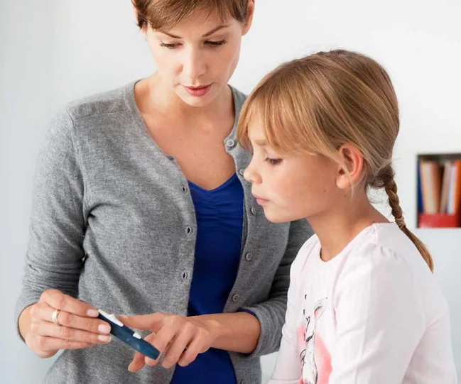 A woman showing a young girl how to use a blood glucose meter in a home setting.