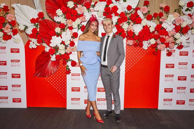 A woman in a blue dress and red hat, and a man in a grey suit with a champagne glass, pose by a floral Colgate sign.