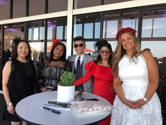 Five people in elegant attire pose smilingly at a table with a potted plant, in a glass-walled venue.