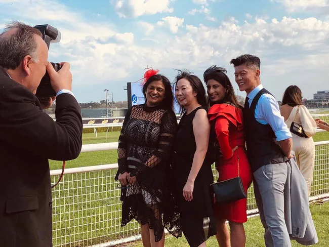 Photographer captures a group of four people dressed up at a racetrack, smiling for a photo in front of a grassy field.