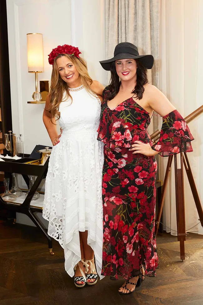 Two women smiling; one in a white dress with a flower headband, the other in a floral dress with a black hat.