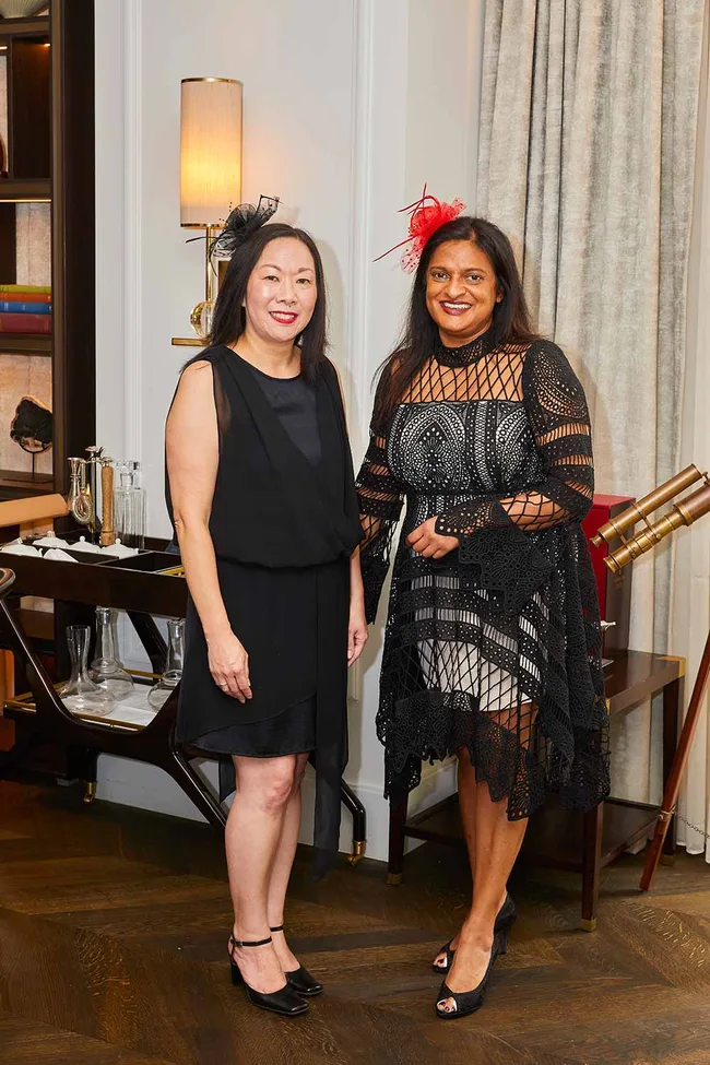Two women in black dresses with fascinators stand smiling in a room with elegant decor.