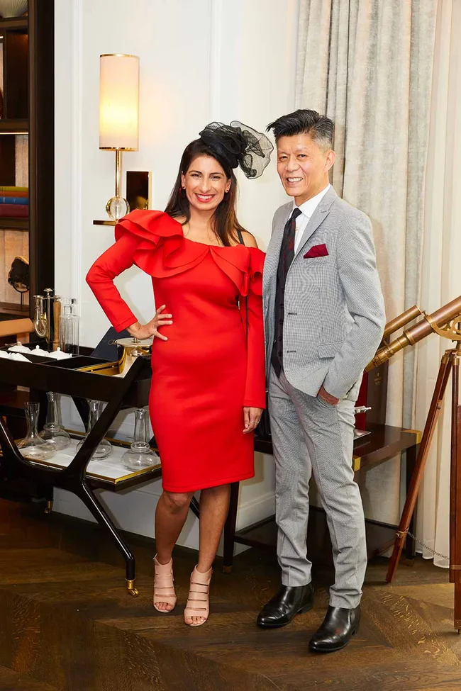 A woman in a red dress and hat poses with a man in a gray suit, standing indoors with elegant decor.