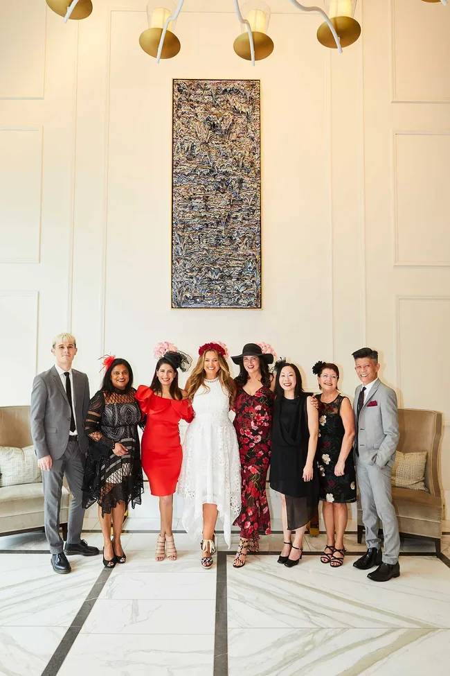 Group of eight people in formal attire, some wearing hats, standing in a decorated room with a wall artwork.