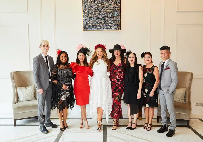 Group of people smiling, dressed in elegant attire with hats, standing in a decorated room.