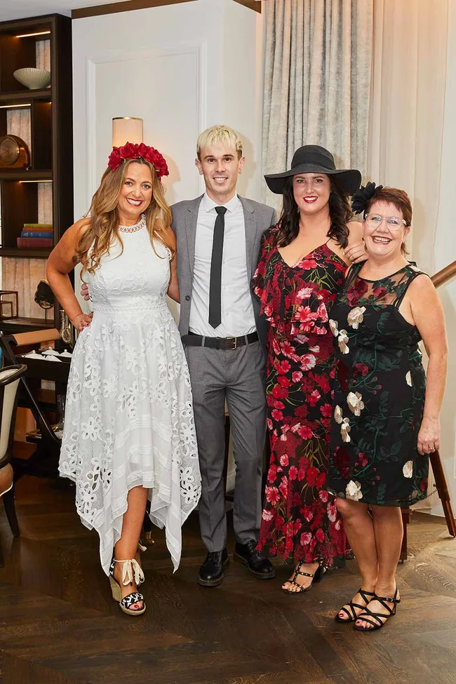 Four people posing; two women in floral dresses, one man in a suit, and another woman in a white dress with red flowers.