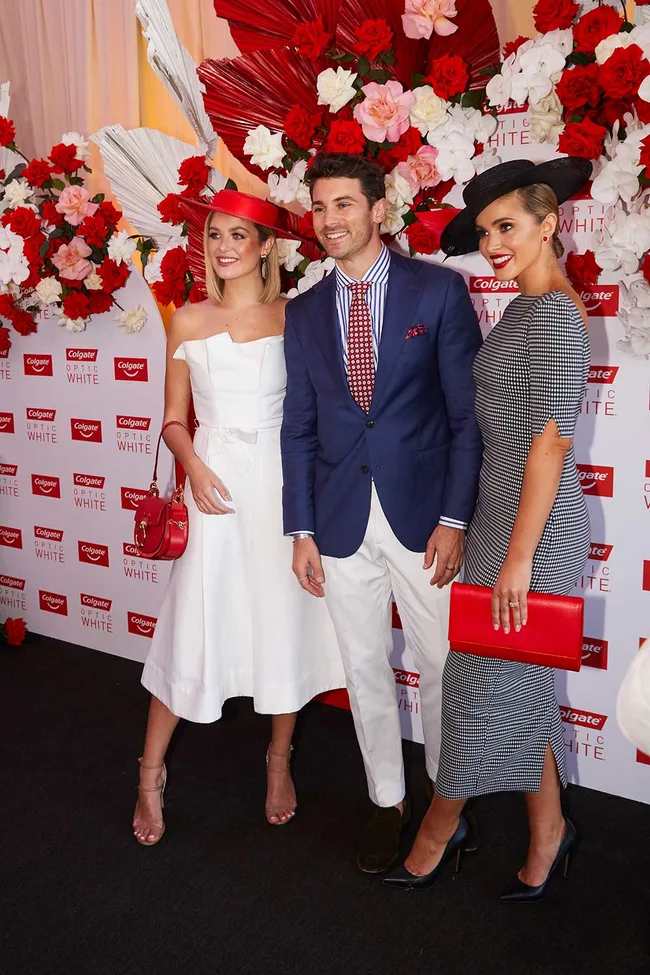 Three people pose together at a Colgate Optic White event, with red and white floral backdrop.
