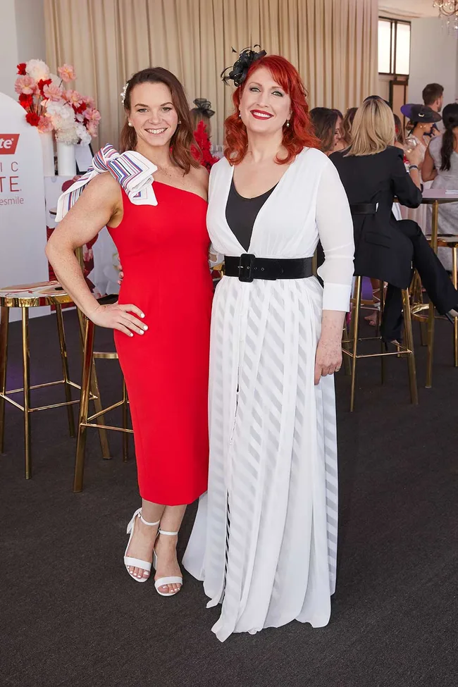 Two women smiling, one in a red dress with a bow, the other in a white dress with a black belt at an indoor event.