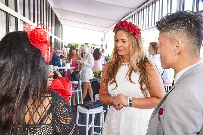 Group of people in formal attire chatting at a lively indoor event; woman wearing a white dress and red floral headband.