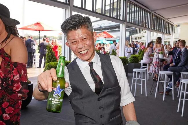 Man in a suit holding a New Zealand Heineken bottle, smiling at a lively indoor event with people in the background.