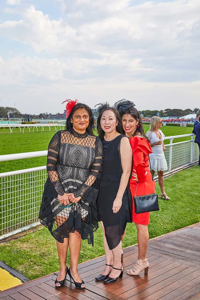 Three women dressed for a horse racing event, posing on a wooden deck with a grass track and railings in the background.