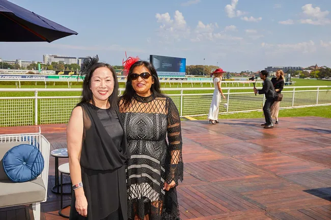 Two women in fashionable dresses and hats pose at a racetrack event under a clear sky.
