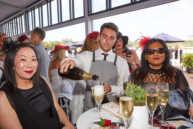 Server pouring champagne at a social event, with two women seated at a table, one wearing sunglasses and a red fascinator.